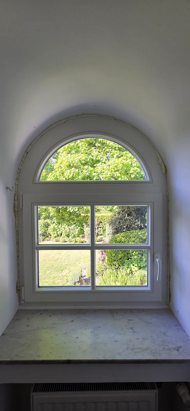 Fenêtre PVC blanche cintrée avec petits-bois, posée dans une baie voûtée, vue sur jardin fleuri