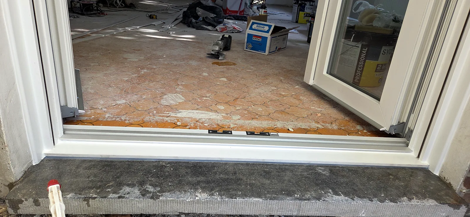 Détail du seuil et des points de verrouillage d'une baie coulissante PVC en cours de pose, avec outils et produits d'étanchéité au sol