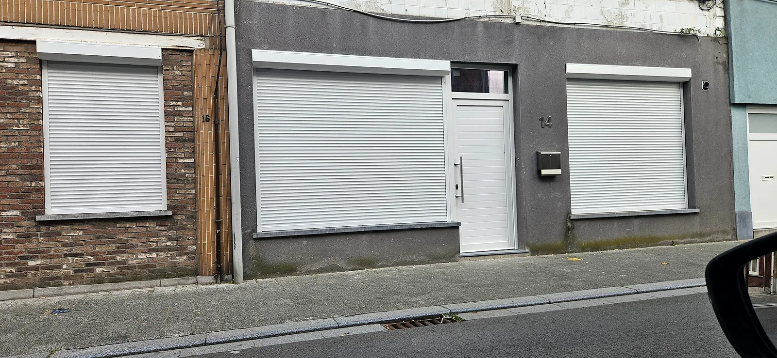Trois volets roulants blancs fermés sur une façade de rue, fenêtres et porte d'entrée